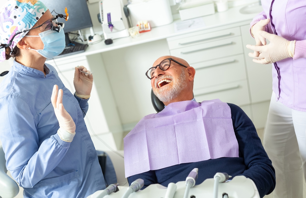 An older man with glasses and a beard laughs while sitting in a dental chair, wearing a lavender bib, as a dentist in scrubs and gloves talks to him in a bright, modern dental office.