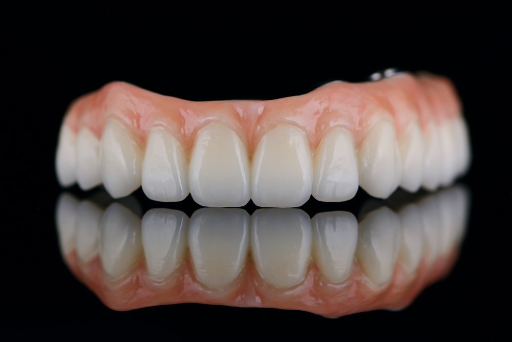 A set of artificial upper teeth with pink gums appears against a black background, with a clear reflection below. The teeth are clean, white, and well-aligned, indicating dental prosthetics or dentures.