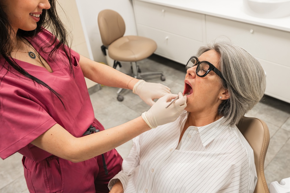 A dental professional in pink scrubs and gloves examines the open mouth of an older woman with gray hair and glasses, who is sitting in a dental chair in a clinic setting.